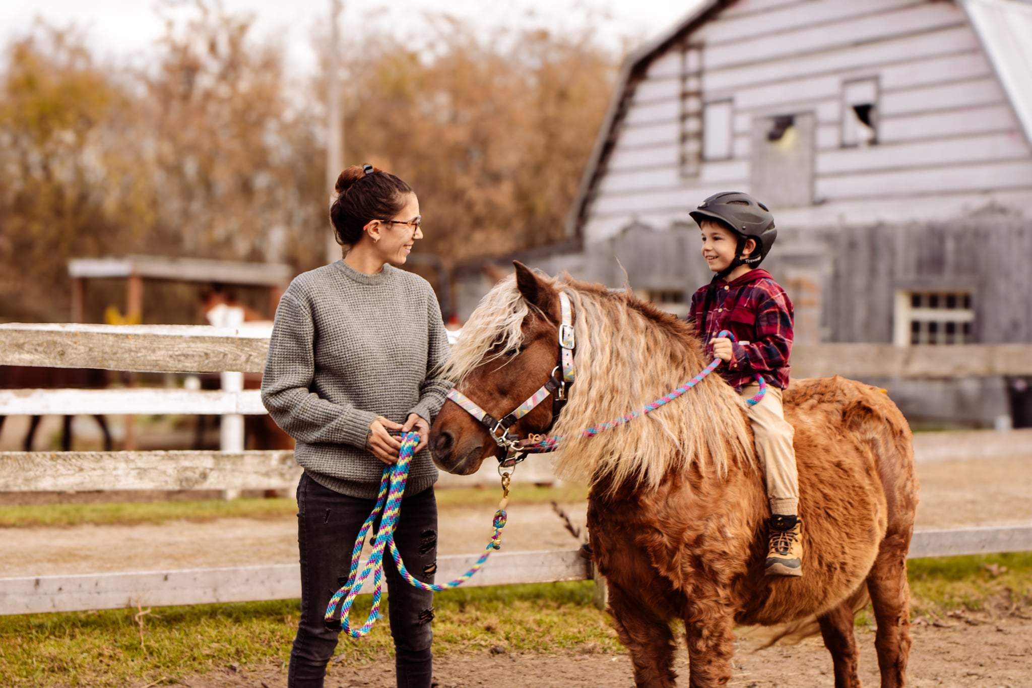Une femme souriante, portant un chandail gris tricoté, tient la bride d'un petit poney brun et hirsute, avec une crinière et une queue claires. Un jeune enfant, portant un casque d'équitation noir et une chemise à carreaux rouge et noir, est assis sur le dos du poney. Ils se regardent tous deux en souriant, avec une vieille grange en bois gris en arrière-plan.