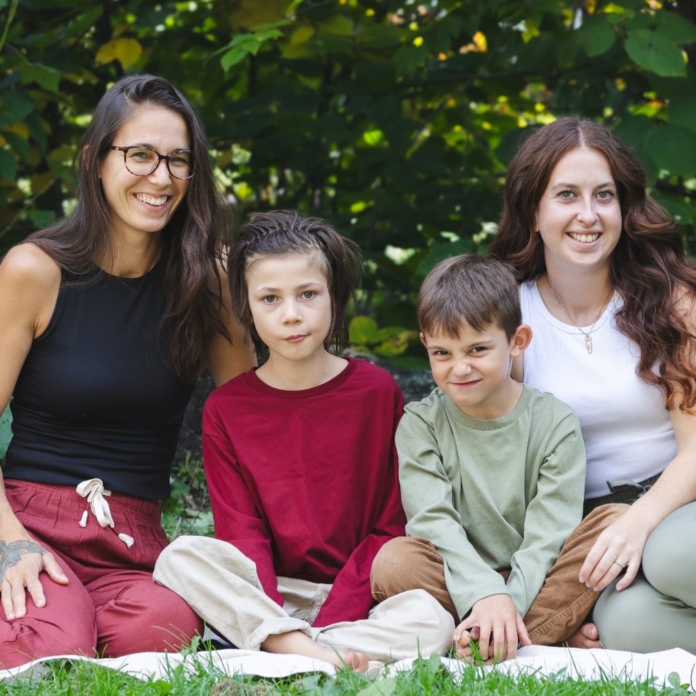 Quatre personnes (deux femmes et deux jeunes garçons) sont assises sur une couverture blanche devant un feuillage dense et vert. Les deux femmes, souriantes, encadrent les garçons. La femme de gauche porte un haut noir sans manches et un pantalon bordeaux, et la femme de droite porte un débardeur blanc et un pantalon vert olive. Les garçons sont vêtus de chemises bordeaux et vert olive. Un bouquet décoratif est visible à l'extrême gauche de l'image.