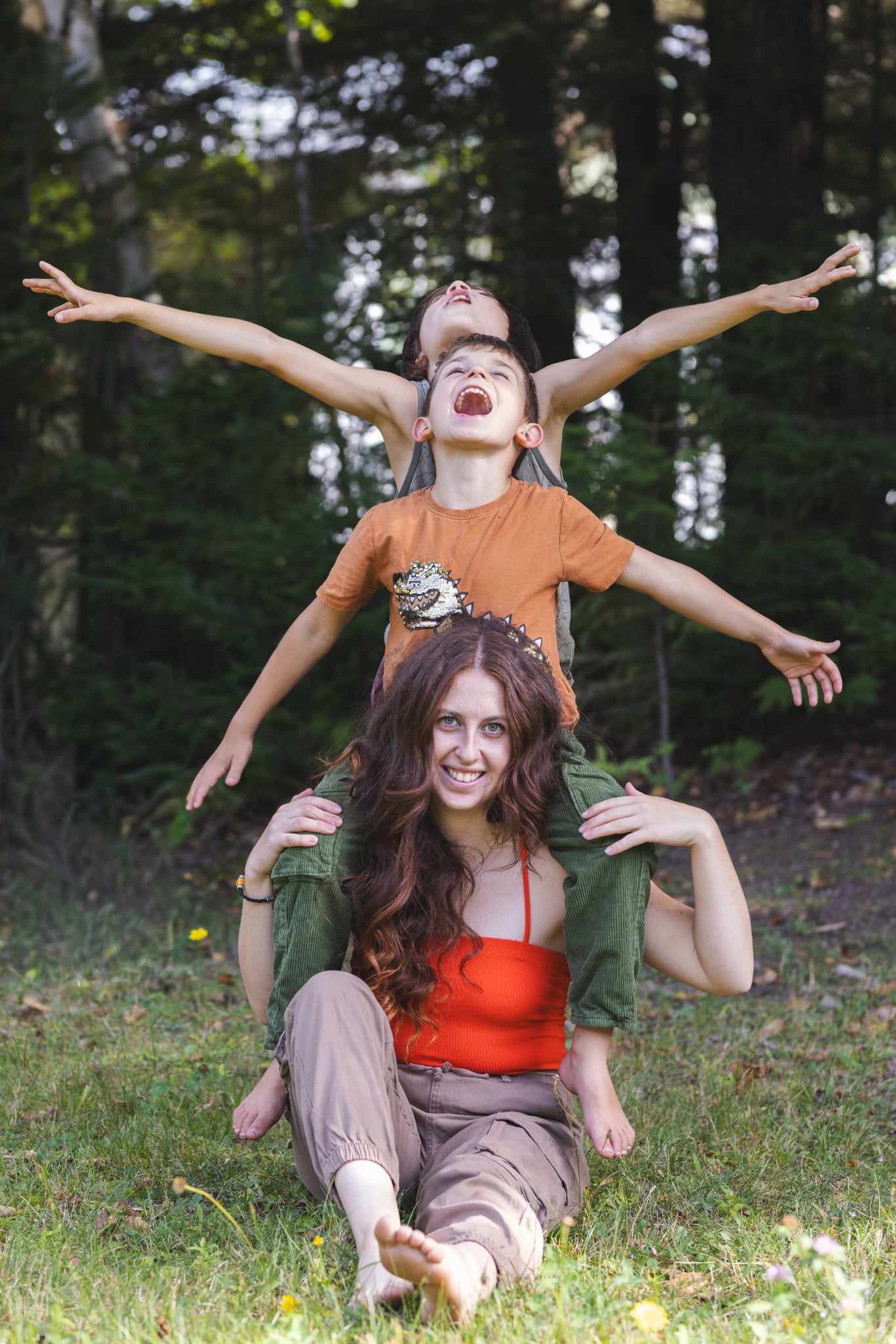 Une femme souriante, assise sur l'herbe avec de longs cheveux bruns et une couronne de feuilles, porte un enfant sur ses épaules. L'enfant, vêtu d'un t-shirt orange, a les bras tendus sur les côtés et la bouche ouverte en signe de joie. Un autre enfant est debout derrière lui, se tenant à l'enfant du milieu, également les bras ouverts. Tous sont devant un dense arrière-plan d'arbres verts.