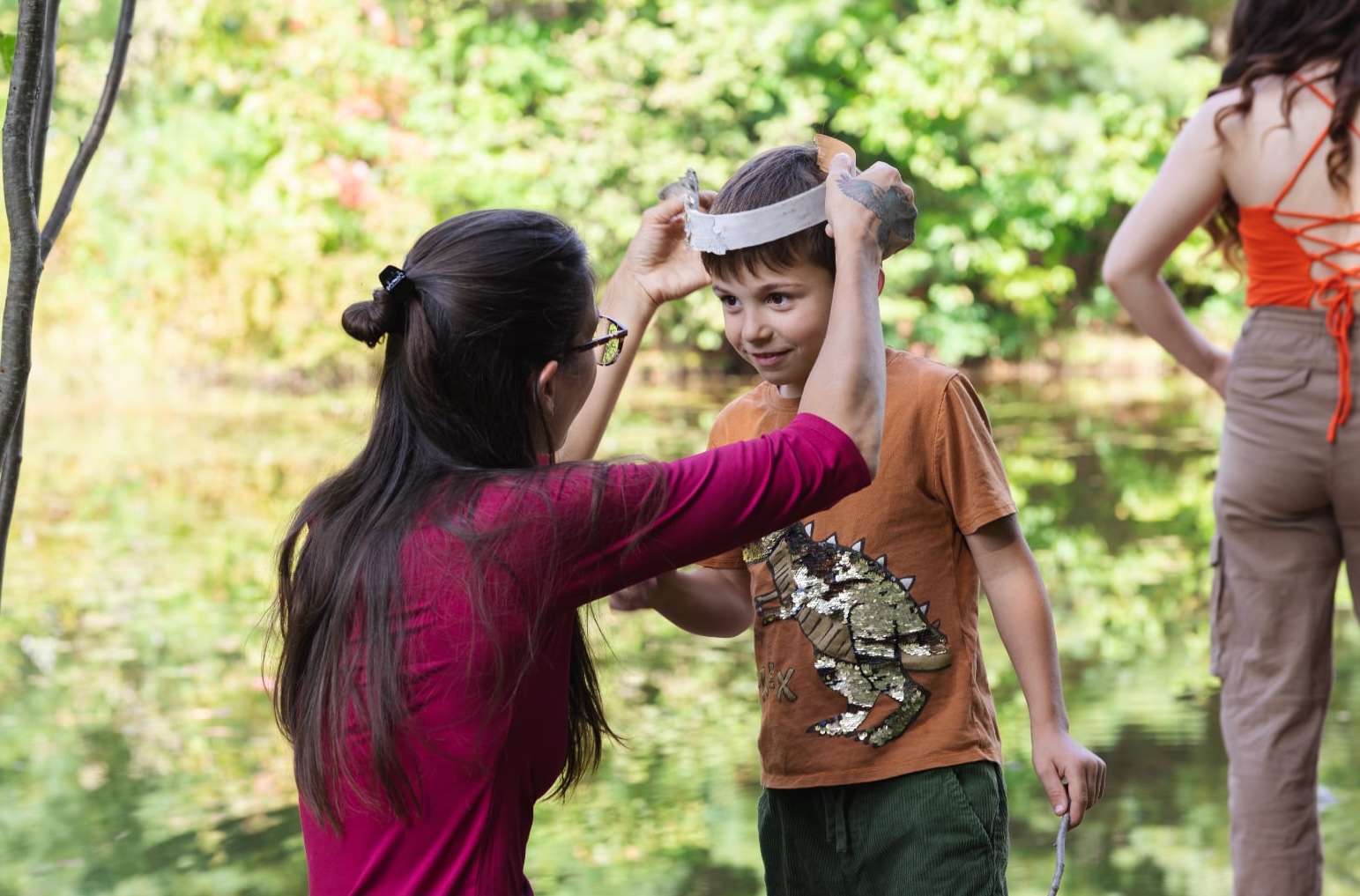 Une femme aux longs cheveux bruns, vêtue d'un haut magenta, est en train de placer un bandeau blanc sur la tête d'un jeune garçon. Le garçon sourit et porte un t-shirt orange avec un motif de dinosaure à paillettes et un pantalon vert. L'arrière-plan est flou, dominé par la verdure et une étendue d'eau. Une autre personne est partiellement visible à droite.